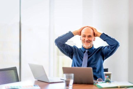 Photo for Shot of mid aged businessman sitting in a modern office and using laptops for work. Professional male wearing shirt and tie. Hand on head. - Royalty Free Image
