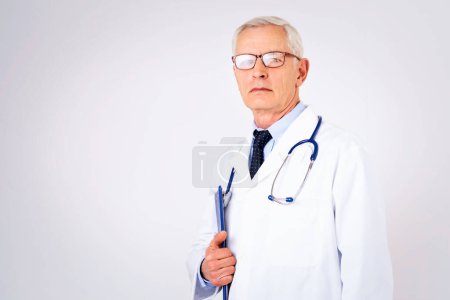 Photo for Senior male doctor wearing lab coat and stethoscope and holding clipboard in his hand while standing at isolated white background. Copy space - Royalty Free Image