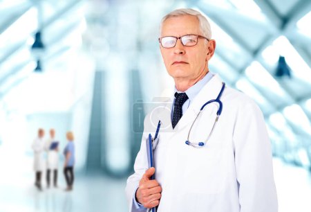 Foto de Médico senior con bata de laboratorio y estetoscopio y sujetando el portapapeles en la mano mientras está parado en el pasillo del hospital - Imagen libre de derechos