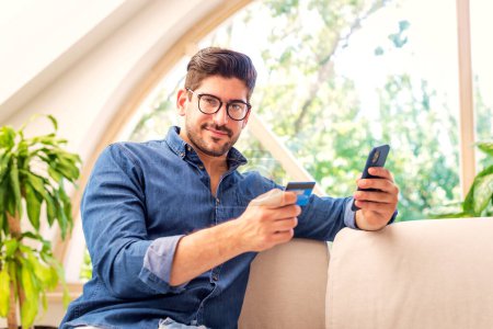 Téléchargez les photos : Prise de vue d'un bel homme assis sur le canapé à la maison et utilisant son téléphone portable et sa carte de crédit pour faire du shopping en ligne. - en image libre de droit