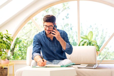 Téléchargez les photos : Portrait d'un homme d'affaires heureux utilisant le téléphone et parlant avec quelqu'un assis sur un canapé et travaillant à la maison. Homme confiant prenant des notes sur le bloc-notes. - en image libre de droit