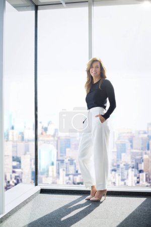 Photo for Full length shot of an attractive businesswoman wearing sweater and trousers while standing at window and looking at camera. - Royalty Free Image