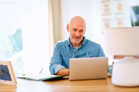 Foto de Un hombre mayor de confianza usando un cuaderno en casa. Hombre de mediana edad trabajando en línea y sentado en el escritorio. Oficina en casa. - Imagen libre de derechos
