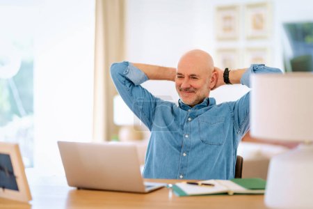 Téléchargez les photos : Homme âgé confiant utilisant un ordinateur portable à la maison. Homme d'âge moyen travaillant en ligne et assis au bureau. Bureau à domicile. Mains derrière la tête. - en image libre de droit