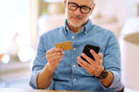 Téléchargez les photos : Homme d'âge moyen assis à la maison et utilisant la carte de crédit et le téléphone mobile pour faire du shopping en ligne. Homme confiant portant chemise et lunettes. - en image libre de droit