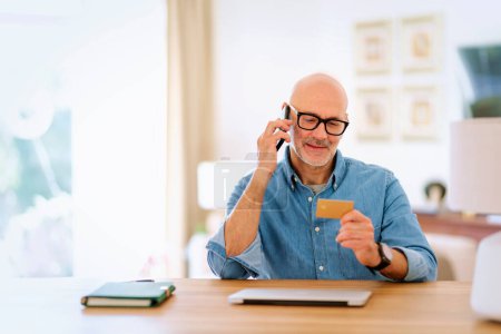 Photo for Mid aged man sitting at home and using credit card and mobile phone for shopping online. Confident male wearing shirt and eyewear. - Royalty Free Image