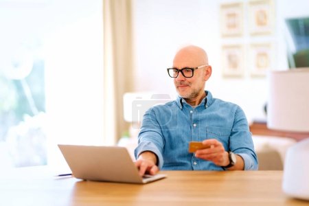 Téléchargez les photos : Homme d'âge moyen assis à la maison et utilisant une carte de crédit et un ordinateur portable pour faire du shopping en ligne. Homme confiant portant chemise et lunettes. - en image libre de droit