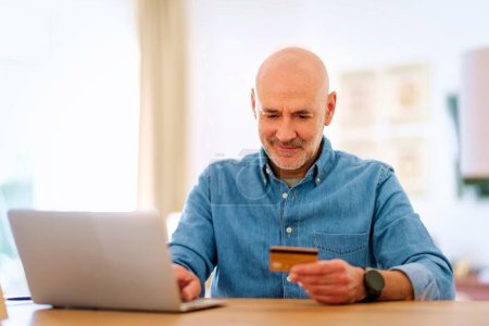 Téléchargez les photos : Homme d'âge moyen assis à la maison et utilisant une carte de crédit et un ordinateur portable pour faire du shopping en ligne. Homme confiant portant chemise et souriant. - en image libre de droit