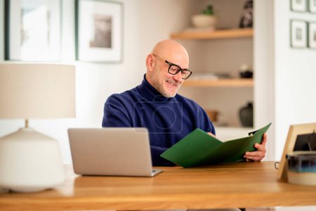 Téléchargez les photos : Heureux homme d'âge moyen en utilisant un ordinateur portable et en prenant des notes tout en travaillant à la maison. Bureau à domicile. - en image libre de droit