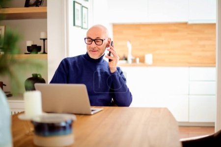 Téléchargez les photos : Confident middle aged man making a call and using laptop while working from home. Confident male sitting in the kitchen and having a call. Home office. - en image libre de droit