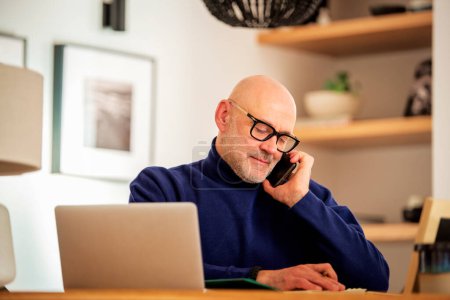 Téléchargez les photos : Confident middle aged man making a call and using laptop while working from home. Confident male sitting in the kitchen and having a call. Home office. - en image libre de droit