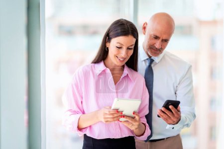 Photo for Shot of businesswoman and businessman standing at the window in the modern office. Professional woman using digital tablet while businessman holding smartphone in his hand. Teamwork. - Royalty Free Image