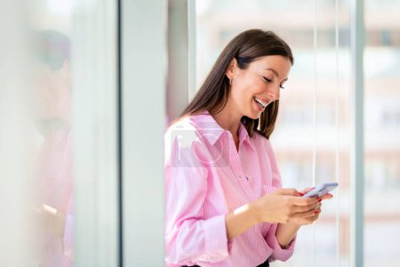 Photo for Brunette haired mid aged businesswoman standing in a modern office and text messaging. Confident professional female wearing shirt and cheerful smiling. - Royalty Free Image