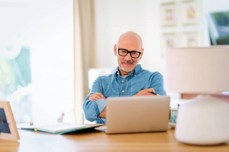 Téléchargez les photos : Homme âgé confiant utilisant un ordinateur portable à la maison. Homme d'âge moyen travaillant en ligne et assis au bureau. Bureau à domicile. - en image libre de droit