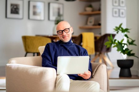 Téléchargez les photos : Homme âgé confiant utilisant un ordinateur portable à la maison. Homme d'âge moyen travaillant en ligne et dans un fauteuil. Bureau à domicile. - en image libre de droit