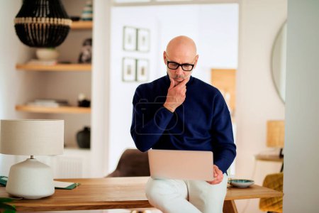 Photo for Confident senior man using notebook at home. Mid aged male working online and sitting at desk. Home office. Professional male looking thoughtfully. - Royalty Free Image