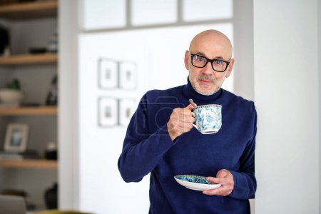 Photo for Portrait of a middle-aged man standing in the apartment drinking tea. Man wearing eyewear and casual clothes. - Royalty Free Image
