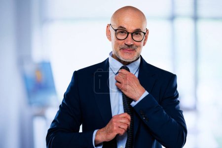 Téléchargez les photos : Un homme d'affaires en costume et cravate debout au bureau. Homme professionnel portant des lunettes et ajustant sa cravate. - en image libre de droit