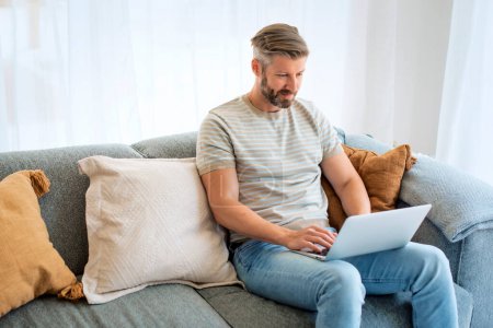Foto de Hombre de mediana edad sentado en el sofá en casa y usng portátil para el trabajo, videollamada. Caucásico masculino usando ropa casual. - Imagen libre de derechos