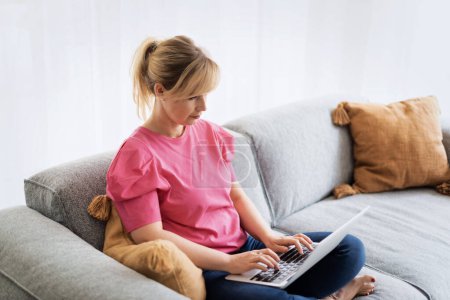 Téléchargez les photos : Femme aux cheveux blonds assise sur un canapé et utilisant un ordinateur portable pour le travail ou ayant un appel vidéo. Femme confiante portant des vêtements décontractés et souriant. Bureau à domicile. - en image libre de droit