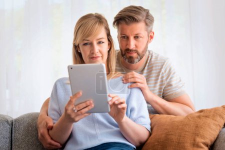 Téléchargez les photos : Joyeux couple relaxant sur le canapé à la maison. Attrayant femme d'âge moyen et bel homme utilisant une tablette numérique et browisng sur Internet ou ayant appel vidéo. - en image libre de droit