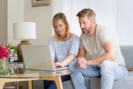 Photo for Happy couple relaxing on the sofa at home. Attractive mid aged woman and handsome man using laptop and browisng on the internet or having video call. - Royalty Free Image