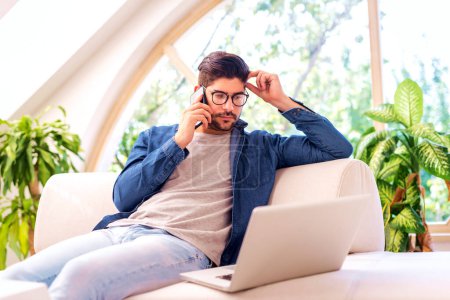 Téléchargez les photos : Homme d'affaires utilisant un ordinateur portable et un téléphone portable à la maison. Jeune homme assis sur le canapé et travaillant à la maison. - en image libre de droit