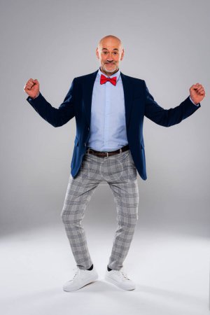 Téléchargez les photos : Studio shot of funny faced man checked pants and bow tie while standing at isolated grey background. Espace de copie. - en image libre de droit