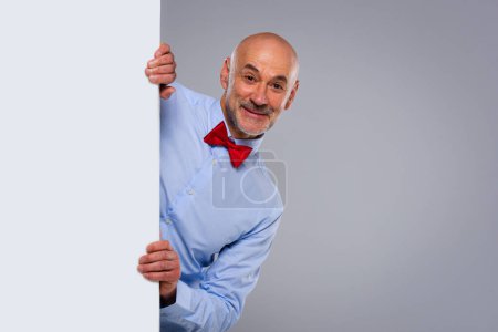 Photo for Studio shot of funny faced middle aged man looking out of white board while standing at isolated grey background. Copy space. - Royalty Free Image