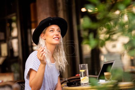 Téléchargez les photos : Jeune femme assise à l'extérieur à la terrasse typique du café français et fumant la cigarette. Chapeau féminin élégant. Street look mode. - en image libre de droit