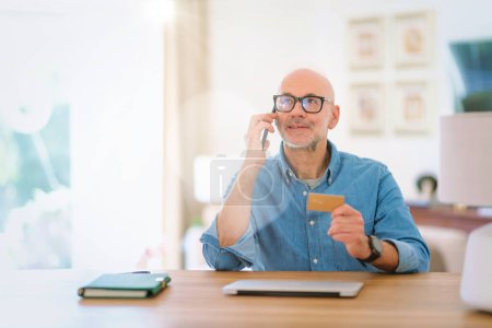 Téléchargez les photos : Homme d'âge moyen assis à la maison et utilisant la carte de crédit et le téléphone mobile pour faire du shopping en ligne. Homme confiant portant chemise et lunettes. - en image libre de droit