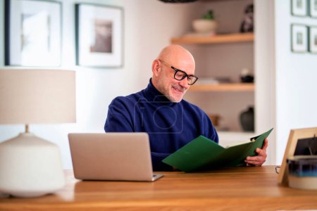 Téléchargez les photos : Homme âgé confiant utilisant un ordinateur portable à la maison. Homme d'âge moyen travaillant en ligne et écrivant quelque chose au bureau. Bureau à domicile. - en image libre de droit