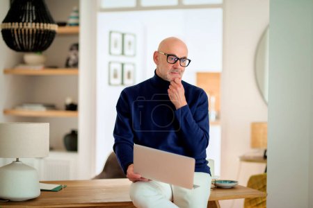 Téléchargez les photos : Homme âgé confiant utilisant un ordinateur portable à la maison. Homme d'âge moyen regardant attentivement tout en travaillant à la maison. Bureau à domicile. - en image libre de droit