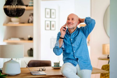 Foto de Tiro de hombre maduro usando el ordenador portátil y hacer una llamada mientras está de pie en el escritorio y trabajando desde casa. - Imagen libre de derechos