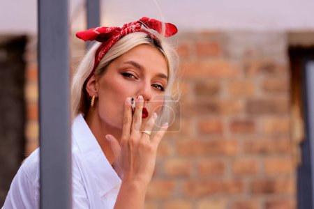 Foto de Primer plano de la hermosa mujer joven que usa bufanda para el cabello y camisa blanca mientras fuma cigarrillos y está de pie en el balcón. - Imagen libre de derechos