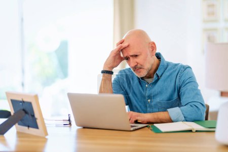 Photo for Worried senior man using notebook at home. Mid aged male working online and sitting at desk. Home office. - Royalty Free Image