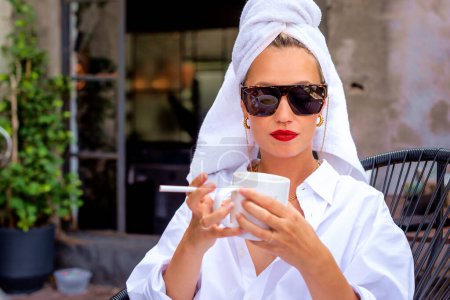 Foto de La joven confiada que usa turbante de toalla es su cabeza y gafas de sol. Atractiva hembra bebiendo café de la mañana y fumando cigarrillo en el balcón. - Imagen libre de derechos