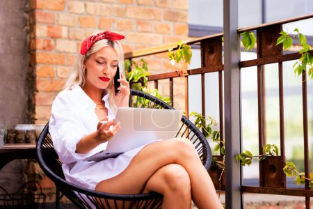 Foto de Mujer atractiva fumando cigarrillo y tener una llamada telefónica mientras trabaja en el ordenador portátil en el balcón en casa. Mujer de pelo rubio con bufanda de cabeza de estilo parisino y camisa blanca de gran tamaño. - Imagen libre de derechos