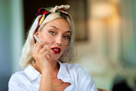 Photo for Clolse-up shot of beautiful young woman wearing hair scarf and white shirt while smoking cigarette. Attractive having blond hair and wearing red lipstick. - Royalty Free Image