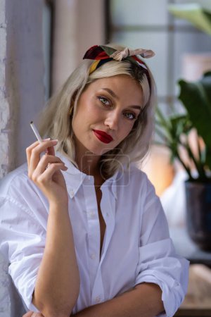 Téléchargez les photos : Portrait d'une belle jeune femme portant une écharpe à cheveux et une chemise blanche tout en fumant une cigarette près de la fenêtre à la maison. - en image libre de droit