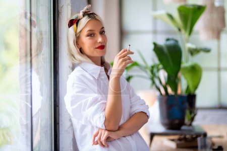 Foto de Retrato de una hermosa mujer joven que usa bufanda para el cabello y camisa blanca mientras fuma cigarrillo junto a la ventana en casa. - Imagen libre de derechos