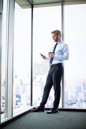Photo for Full length of smiling businessman using smartphone and earphones while standing at the office. Professional man having video call and drinking coffee. - Royalty Free Image