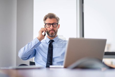 Téléchargez les photos : Homme d'affaires sérieux face assis au bureau et à l'aide d'un ordinateur portable et téléphone mobile pour le travail. professionnel masculin d'âge moyen ayant un appel d'affaires. - en image libre de droit