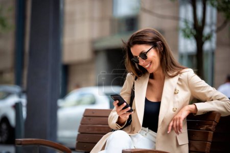 Photo for Attractive brown-haired woman is sitting on a bench in the street and using phone. Cheerful smiling female wearing business casual and text messaging. - Royalty Free Image