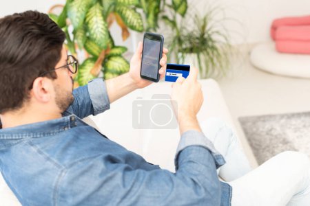Photo for Rear view of an unrecognizable man is sitting at home holding his mobile phone and bank card in his hand. Man banking online. - Royalty Free Image