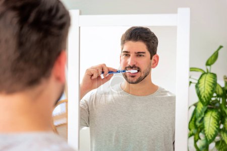 Foto de Un hombre con cara de rastrojo de pie en el baño delante del espejo y cepillándose los dientes. - Imagen libre de derechos