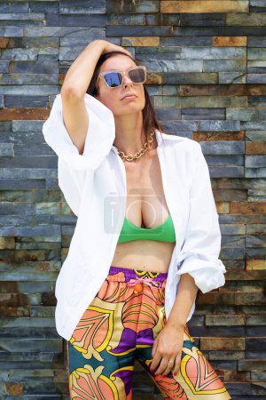 Téléchargez les photos : Portrait d'une jolie femme portant des lunettes de soleil et des vêtements de plage et se relaxant au mur. - en image libre de droit