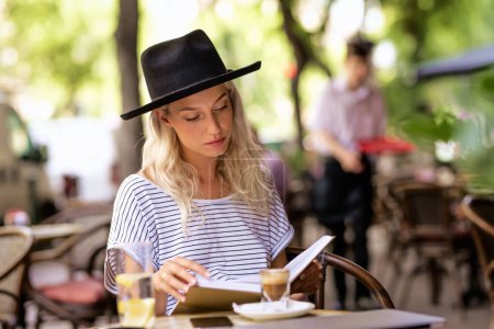 Téléchargez les photos : Une femme aux cheveux blonds et au chapeau est assise sur la terrasse d'un café et lit un livre. - en image libre de droit