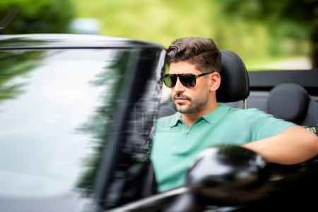 Photo for A handsome man wearing sunglasses and sitting in a convertible car and driving on the road. - Royalty Free Image