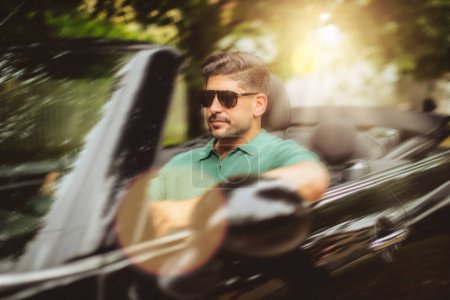 Foto de Un hombre guapo con gafas de sol y sentado en un coche convertible y conduciendo en la carretera. - Imagen libre de derechos